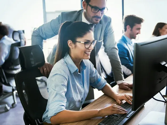 Smiling employee typing at a desktop as a manager looks on, while colleagues work at desks in an open office